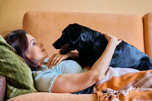 Relaxed middle aged woman lying on sofa under blanket and petting black labrador puppy dog. Lifestyle, love, care, pets, 40s people concept