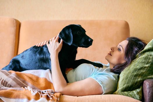 Relaxed middle aged woman lying on sofa under blanket and petting black labrador puppy dog. Lifestyle, love, care, pets, 40s people concept