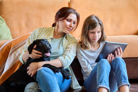 Family, lifestyle, communication, happiness, vacation together, pets, home concept. Mother daughter child and labrador puppy dog sitting together near sofa looking into digital tablet screen at home