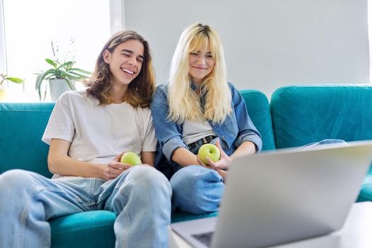 Laughing couple of teenagers having fun, sitting on couch, looking at laptop screen, with green apples in hands. Teens, youth, friendship, lifestyle, leisure concept