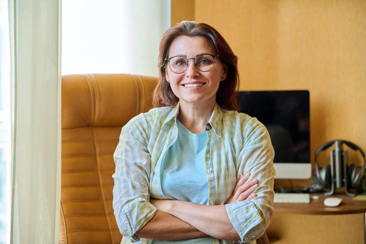 Portrait of confident smiling middle aged woman looking at camera with crossed arms at home workplace, 40s female with glasses sitting on armchair near computer