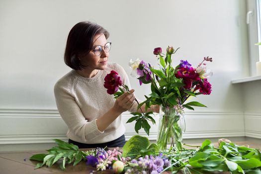 Mature woman at home with spring flowers. Female making bouquet, placing flowers in jug of water. Spring summer season, floristry, home decoration, natural beauty concept