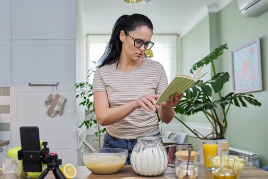 Woman preparing apple pie at home in kitchen with recipe book in her hand, looking in smartphone webcam, talking online using video call. Female food blogger recording video recipe