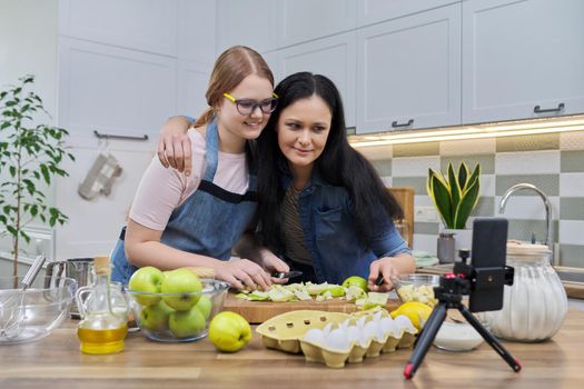 Mom and teen daughter cooking apple pie together, looking at smartphone screen, video recipe, writing recipe on family food blog, talking online using video communication