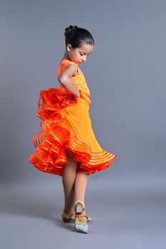 Child girl in an orange sports dress posing in dance movement on gray studio background, sports dances ballroom and latin american for children and adolescents