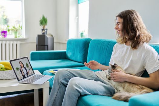 Guy talking online with teacher, psychologist, social worker. Teenager sitting at home with pet cat, using video call on laptop, virtual meeting with counselor. Technology, adolescence, mental health