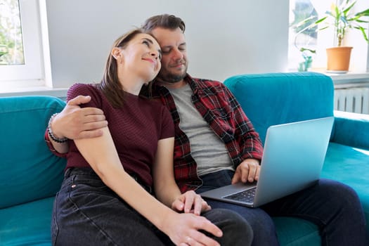 Happy middle aged couple sitting on couch with laptop. Man and woman embracing together looking at laptop screen, in living room. Relationships, lifestyle, communication, home life, middle-aged people