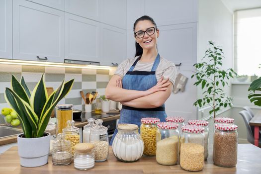 Portrait of smiling woman housewife in an apron in kitchen. Jars of cereals on the table, organizing and storing food