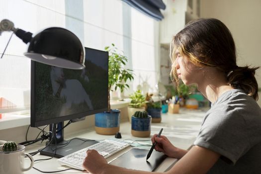 Teen guy sitting at table at home working on computer, using graphics tablet to work with images. Modern technologies, hobby of male teenager, freelance, youth concept