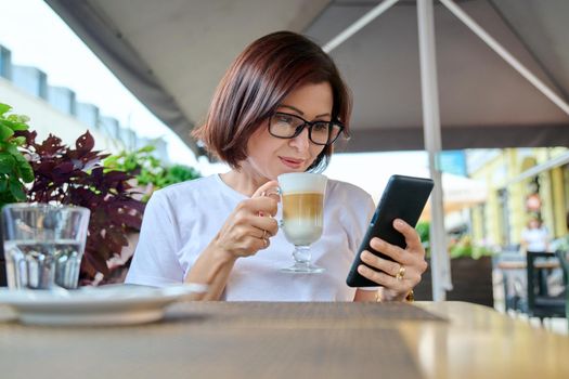 Serious middle-aged woman sitting in an outdoor cafe with a cup of coffee and looking at the smartphone screen. Coffee break, people 40s age, urban style, leisure, lifestyle, technology concept