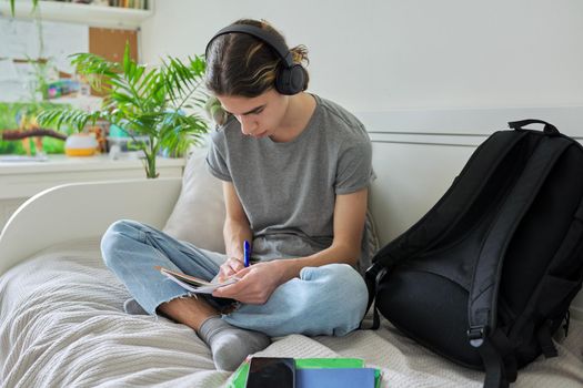 Male teenager with pet green parrot quaker on his shoulder, guy student sitting in headphones with textbooks notebooks books at home on bed