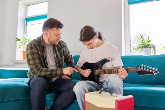 Father teaches his teenage son to play the electric guitar. Family sitting together at home in living room on couch. Parent-child relationship, lifestyle, creativity, music, teenagers concept