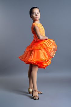 Kids sport dancing, kid girl in orange sport dress posing in dance pose looking at camera on gray studio background.