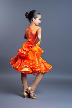 Child girl in an orange sports dress posing in dance movement on gray studio background, sports dances ballroom and latin american for children and adolescents