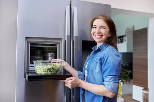 Woman with bowl of vegetable salad from refrigerator. Kitchen interior, open modern chrome refrigerator door background. Eating at home, lifestyle, household, dieting, healthly food concept
