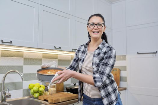 Smiling woman in kitchen mittens with hot freshly baked pie, kitchen interior background