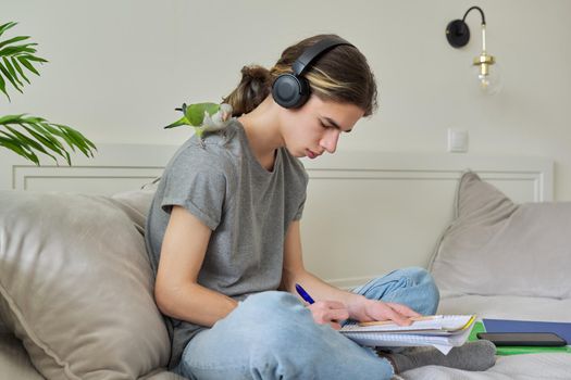 Male teenager with pet green parrot quaker on his shoulder, guy student sitting in headphones with textbooks notebooks books at home on bed