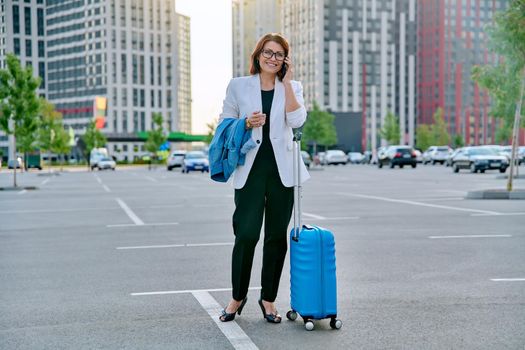 Middle aged businesswoman with suitcase talking on mobile phone in outdoor parking, urban style, city architecture background. Business travel trip, business people of mature age, success, modern city