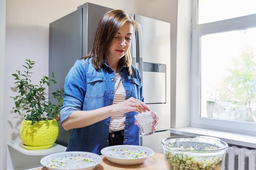 Woman in kitchen near modern chrome large refrigerator with ice for cooling food, vegetable salad in bowl on table. Eating at home, lifestyle, household, dieting, healthly food concept