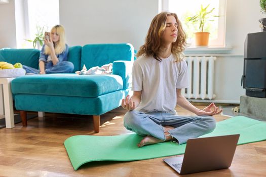 Guy teenager sitting in lotus position on yoga mat at home on floor with laptop. Meditation, online training, leisure, lifestyle, teenagers concept