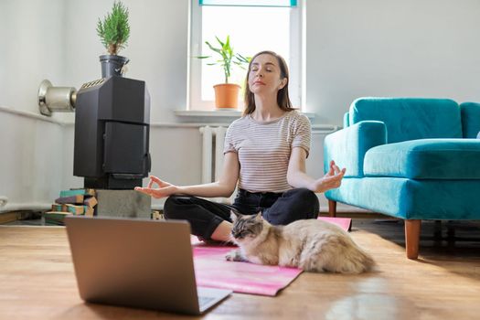 Lifestyle leisure meditation. Middle aged woman sitting at home on floor with laptop in lotus position, pet cat on exercise mat with owner
