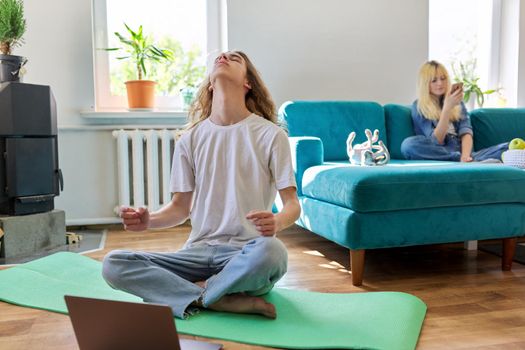 Guy teenager sitting in lotus position on yoga mat at home on floor with laptop. Meditation, online training, leisure, lifestyle, teenagers concept