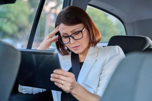 Middle-aged business woman in car in passenger backseat. Serious tired with closed eyes female in glasses with digital tablet. Business people of mature age, city, emotion fatigue meditation sadness