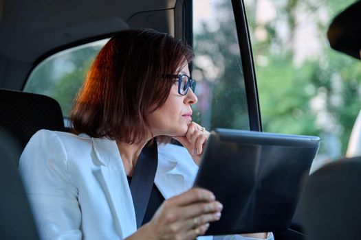 Middle-aged businesswoman in car in passenger backseat. Serious female in glasses with digital tablet, tired woman looking out window. Business people of mature age, city, fatigue frustration