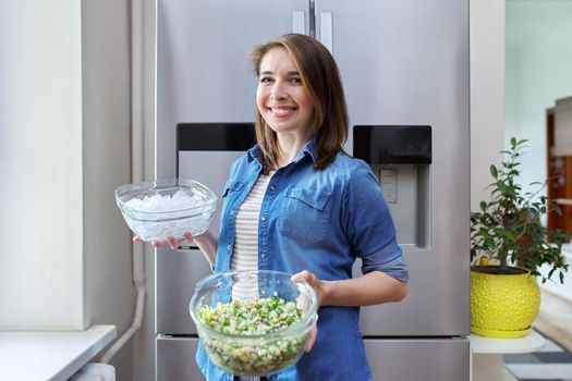 Smiling woman with bowl of ice and plate of vegetable salad in her hands, modern chrome large refrigerator in kitchen. Eating at home, lifestyle, household, dieting, healthly food concept