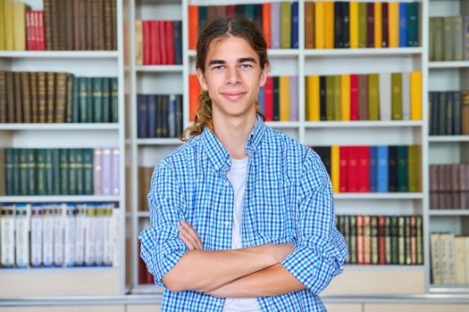 Single portrait of smiling confident male student teenager with crossed arms looking at camera in the library. School, education, knowledge, adolescence concept