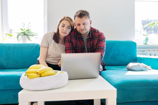 Serious middle-aged couple husband and wife with laptop sitting at home on couch. Man and woman attentively listening and looking at computer monitor, online meeting, video consultation, chat conference