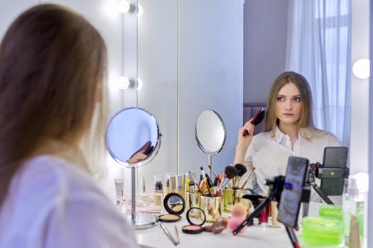 Young beautiful woman combing hair in front of mirror, blonde with long healthy straight hair. Beauty blogger recording video on smartphone, online course, remote training in makeup and hair styling