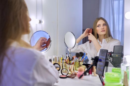 Young beautiful woman combing hair in front of mirror, blonde with long healthy straight hair. Beauty blogger recording video on smartphone, online course, remote training in makeup and hair styling