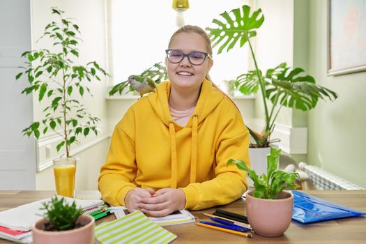 Teenage girl and pet green quaker parrot on the shoulder. Young female student doing school lessons at home in the company of a friend's bird