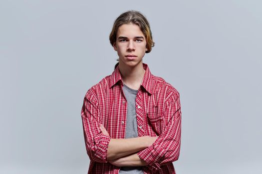 Portrait of serious guy teenager with crossed arms, hipster teen male 15, 16 years old with long hair looking at camera on light background