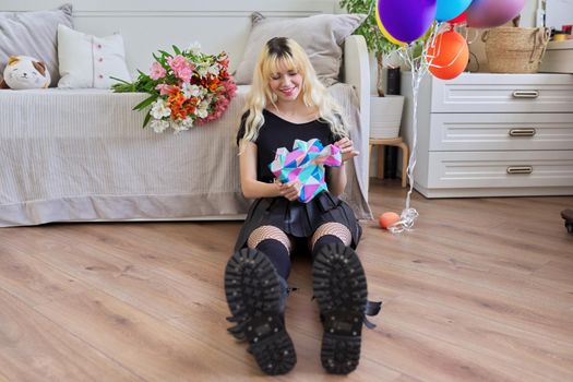 Birthday of teenage female 16, 17 years old. Hipster teenager at home with surprise gift in box, bouquet of flowers, colored balloons, sitting at home on bed. Teens, age, holiday concept