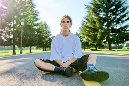 Fashion portrait of hipster teenager guy sitting on road. Serious posing young male with long hair looking in camera,in white t-shirt shorts boots on summer day. Youth, adolescence, people, lifestyle