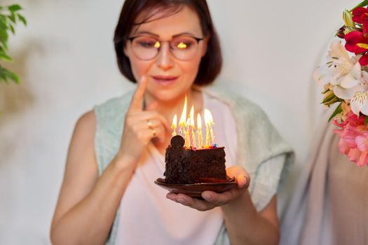 Mature beautiful woman with small birthday cake with burning candles, middle-aged female dreamily making wish and blowing on candles. Woman sitting at home on floor with bouquet of flowers