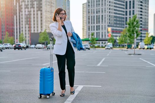 Middle aged businesswoman with suitcase talking on mobile phone in outdoor parking, urban style, city architecture background. Business travel trip, business people of mature age, success, modern city