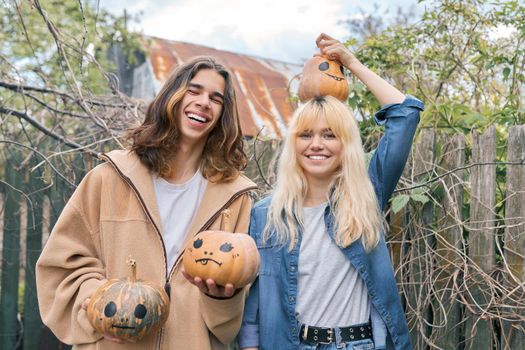 Couple of laughing teenagers with pumpkins having fun outdoors in the garden, halloween autumn holiday, youth concept