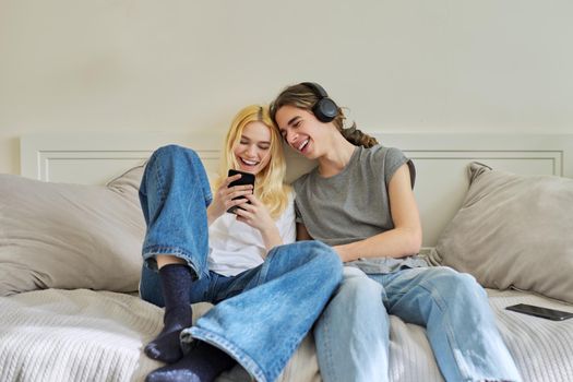 Happy laughing hipster teenagers male and female having fun using smartphone together. Couple using video call, watch funny photos videos. Lifestyle, technology, friendship, communication, adolescents