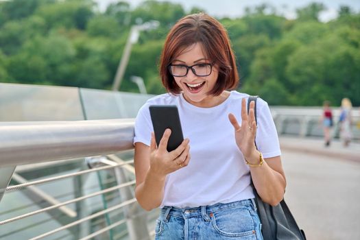 Surprised woman looking in smartphone screen. Middle-aged female with emotion of surprise with phone in her hands on street of summer city