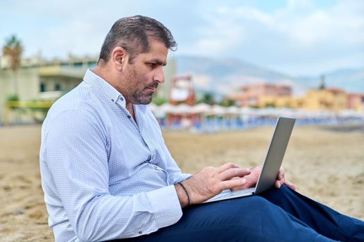 Serious confident mature man with laptop outdoors. Business male working with laptop sitting on sand at beach. Remote business, technology, freelance, lifestyle, leisure, middle age concept