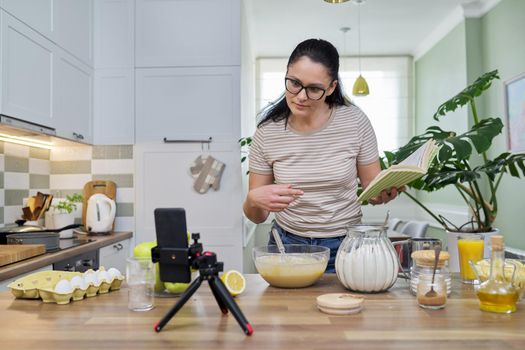 Woman preparing apple pie at home in kitchen with recipe book in her hand, looking in smartphone webcam, talking online using video call. Female food blogger recording video recipe