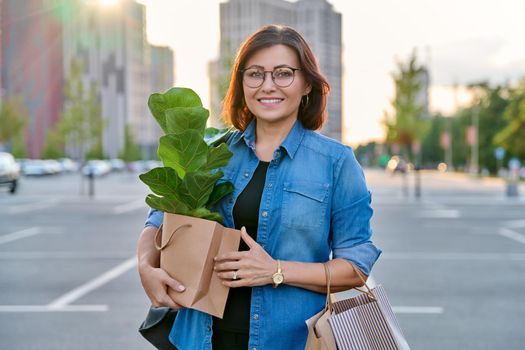 Middle aged woman with paper shopping bags with buying plant, outdoor, store parking lot background. Smiling female shopping in city using craft recycled brown bags, ficus lirata in hand, green trends