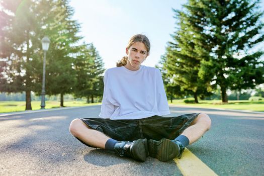 Fashion portrait of hipster teenager guy sitting on road. Serious posing young male with long hair looking in camera,in white t-shirt shorts boots on summer day. Youth, adolescence, people, lifestyle