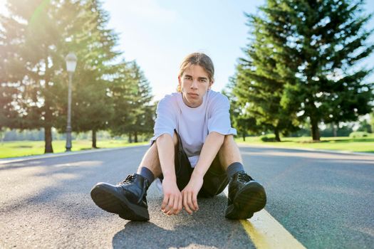 Fashion portrait of hipster teenager guy sitting on road. Serious posing young male with long hair looking in camera,in white t-shirt shorts boots on summer day. Youth, adolescence, people, lifestyle