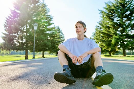 Fashion portrait of hipster teenager guy sitting on road. Serious posing young male with long hair wearing white t-shirt shorts boots on sunny summer day. Youth, adolescence, people, lifestyle concept