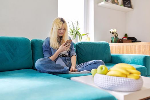 Smiling happy female teenager 16 years old sitting at home on couch looking at smartphone screen, texting, browsing blog, social networks, chatting with friends online