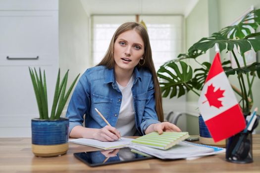 Canada, education, study, students concept. Online training, distance lessons, e-learning, female teenager university student sitting at home looking at webcam, close-up face, Canadian flag on table